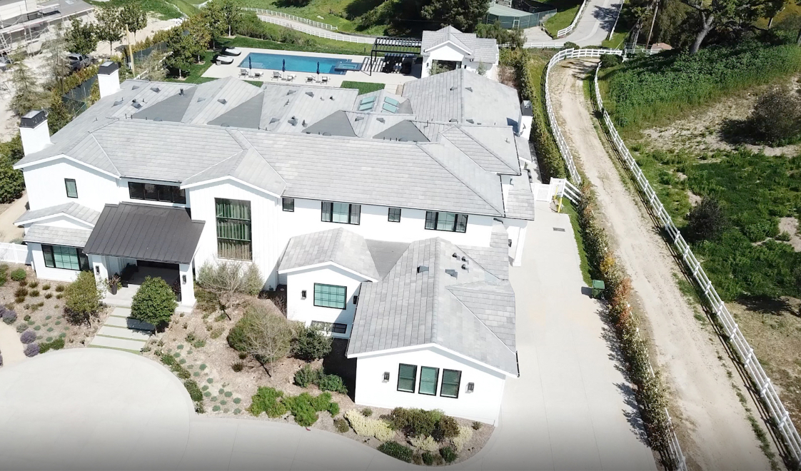 Aerial view of a large white home with a grey roof, an in-ground pool in the backyard, and a pool house